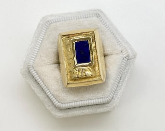 May include: A gold ring featuring a rectangular, dark blue stone. The ring has a textured, rectangular design around the stone. It is presented in a grey, hexagonal ring box. The ring's design is classic.