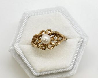 May include: A gold ring featuring a central pearl and two small diamonds. The ring has an ornate, open design with a twisted rope pattern. The ring is presented in a light grey hexagonal ring box.