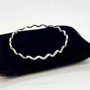 May include: A silver-colored bangle bracelet with a wavy, textured design. The bracelet is circular and rests on a dark brown velvet pillow against a white background. The bracelet's design is reminiscent of ocean waves.
