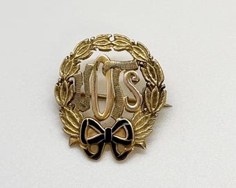 Puede incluir: Broche dorado con las letras "UJS" en el centro, rodeado por una corona de laurel. Un lazo negro y dorado en la parte inferior. El broche es probablemente vintage o antiguo.