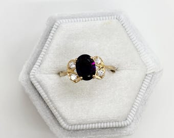 May include: A gold ring featuring an oval, dark purple gemstone. The ring is adorned with small, clear gemstones flanking the main stone. It's presented in a light grey, hexagonal ring box. The ring's design is elegant.