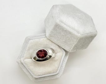 May include: A silver ring with a red gemstone set in a bezel setting. The ring is in a white velvet ring box.