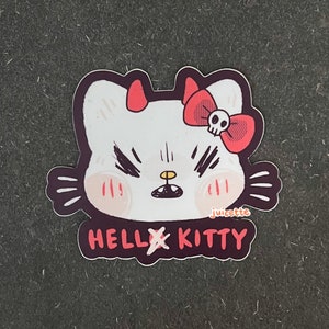 May include: A white Hello Kitty sticker with red devil horns and a red bow with a skull. The text "HELLxKITTY" is below the image. The sticker is on a dark grey background.