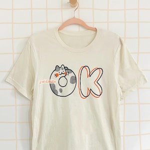 May include: A white t-shirt with a graphic of a cat inside a donut with the word "juicette" and the letter "K" in orange.