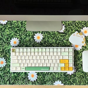 May include: A green and white desk mat with a daisy pattern. The mat features a white keyboard with yellow keys, a white mouse, a white cat-shaped mousepad, and a white mug with a strawberry design. The text "JOYCE HWANG" is printed in the bottom right corner of the mat.