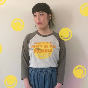 Puede incluir: Una camiseta de béisbol gris y verde con el texto "When in doubt, don't be an asshole" en letras naranja y amarillas. La camiseta presenta una gran cara sonriente amarilla con un contorno naranja.