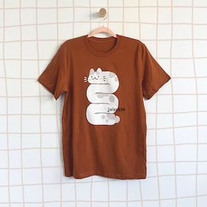 May include: A brown t-shirt with a white graphic of a cat curled up in a spiral shape. The cat has black eyes and a pink nose. The text "juicette" is printed below the cat.