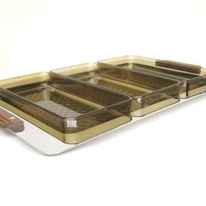 Puede incluir: Una bandeja de servir con tres compartimentos rectangulares de color &aacute;mbar. Los compartimentos tienen un patr&oacute;n texturizado y est&aacute;n colocados sobre una base de acero inoxidable. Las asas de madera est&aacute;n unidas a la bandeja y a los compartimentos.