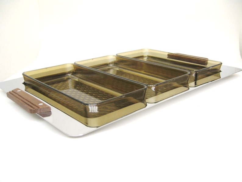 Puede incluir: Una bandeja de servir con tres compartimentos rectangulares de color &aacute;mbar. Los compartimentos tienen un patr&oacute;n texturizado y est&aacute;n colocados sobre una base de acero inoxidable. Las asas de madera est&aacute;n unidas a la bandeja y a los compartimentos.