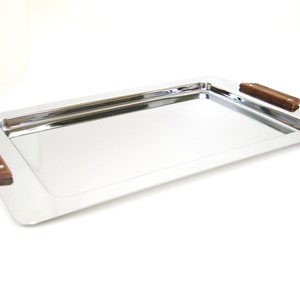 Puede incluir: Una bandeja rectangular para servir con una superficie de acero inoxidable pulido y reflectante. La bandeja tiene dos asas rectangulares de madera marr&oacute;n oscuro en cada extremo. La bandeja est&aacute; dise&ntilde;ada para servir alimentos y bebidas.