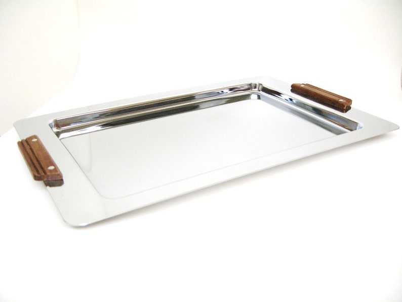 Puede incluir: Una bandeja rectangular para servir con una superficie de acero inoxidable pulido y reflectante. La bandeja tiene dos asas rectangulares de madera marr&oacute;n oscuro en cada extremo. La bandeja est&aacute; dise&ntilde;ada para servir alimentos y bebidas.