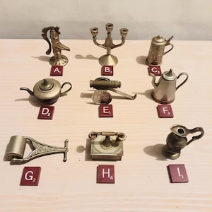 Peut inclure: Ensemble de petits objets en laiton et métal, dont un chandelier, une théière, une chope à bière et un canon. Les articles sont disposés sur une surface claire, chacun avec une petite tuile rouge marquée d'une lettre et d'un chiffre.