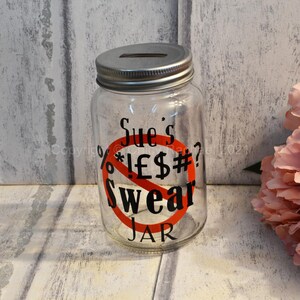 May include: A clear glass jar with a silver lid and a red circle with a diagonal line through it. The jar has the text "Sue's Swear Jar" printed on it.