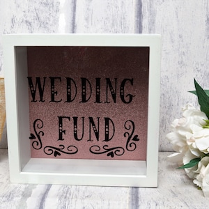 May include: White wooden box with a pink glitter background and black lettering that says "Wedding Fund". The box has a decorative black swirl design at the bottom.
