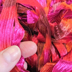 May include: Close-up of vibrant, textured ribbon in shades of pink, red, and orange. The ribbon appears to be made of a shiny, possibly silk-like material. The image shows a hand holding a section of the ribbon, highlighting its texture and color variations.
