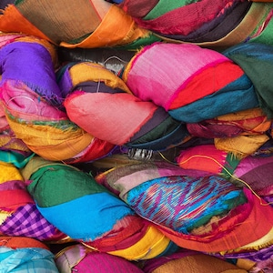 May include: A close-up of a pile of colorful fabric scraps. The fabric pieces are in various colors, including pink, orange, purple, green, and blue. The edges of the fabric are frayed, and the pieces are arranged in a tangled, layered pattern. The image is brightly lit.