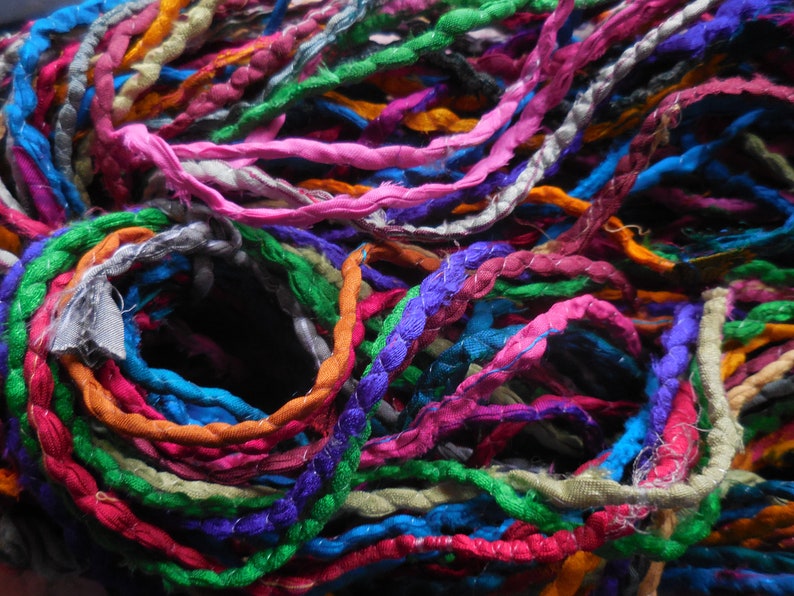 May include: A close-up of a bundle of colorful fabric yarn. The yarn is made of various colors, including pink, blue, green, orange, and purple. The yarn is twisted and knotted together, creating a textured and intricate pattern.