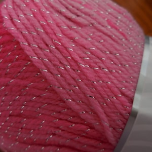 Puede incluir: Hilo rosa con hilo de purpurina plateado. El hilo est&aacute; enrollado en una bola y est&aacute; parcialmente envuelto en una etiqueta blanca.