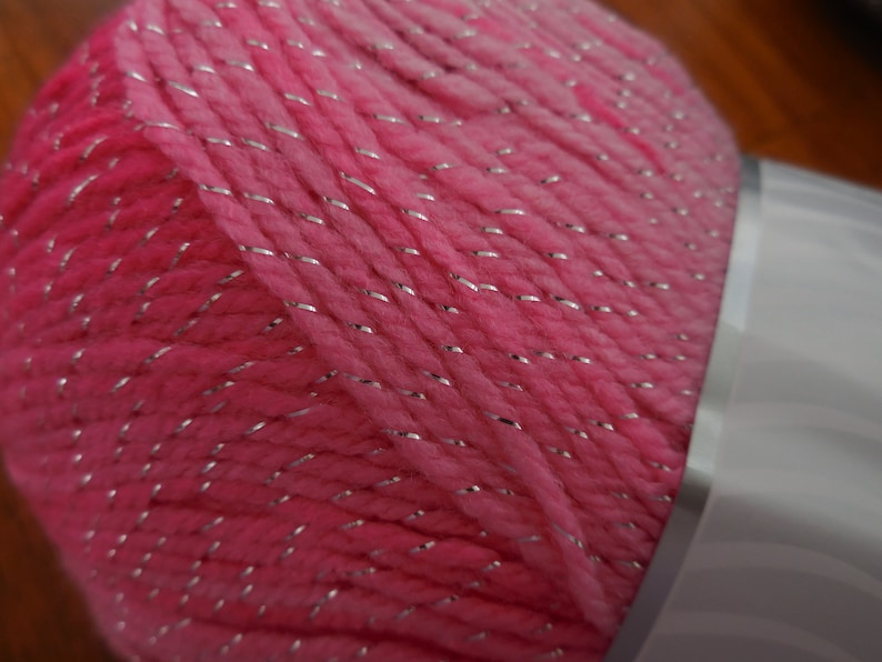 Puede incluir: Hilo rosa con hilo de purpurina plateado. El hilo est&aacute; enrollado en una bola y est&aacute; parcialmente envuelto en una etiqueta blanca.