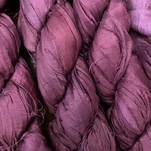 May include: A bundle of deep plum-colored silk ribbon yarn featuring a highly textured, crinkled surface with visible frayed edges and a matte finish. The material is twisted into thick, overlapping strands that create a dense, layered appearance.