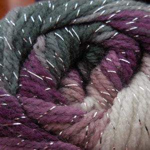 May include: A skein of yarn with a variegated color pattern of purple, gray, and white with silver metallic threads woven throughout.