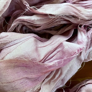 May include: Close-up of a pile of light purple silk ribbon. The ribbon has a soft, flowing texture with frayed edges, creating a delicate and textured appearance. The color varies from pale lavender to deeper shades of purple.