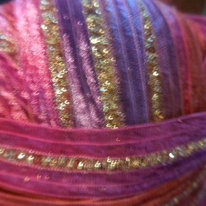 May include: A head covering featuring a vibrant mix of pink, purple, and orange fabric strips arranged in a pleated pattern. The design is accented with thin, shimmering gold sequin bands that run vertically across the textured material.