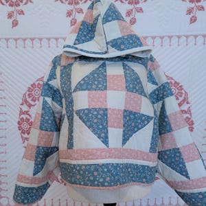May include: A hooded pullover jacket constructed from quilted fabric featuring a patchwork design. The pattern consists of alternating squares and triangles in dusty blue with small floral prints, soft pink with tiny white dots, and solid cream.