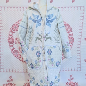 May include: A long-sleeved, hooded quilted coat in off-white fabric featuring a cross-stitch pattern of blue birds, green deer, and floral motifs in blue, pink, and green. The coat includes two large front pockets and a front button closure.