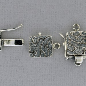 May include: Silver square clasp with a textured design. The clasp has a spring-loaded mechanism and a loop for attaching to a necklace or bracelet.