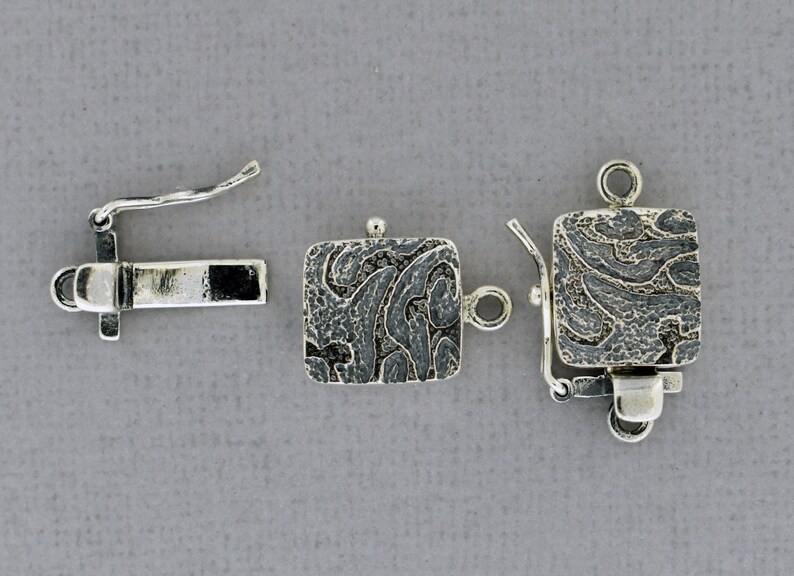 May include: Silver square clasp with a textured design. The clasp has a spring-loaded mechanism and a loop for attaching to a necklace or bracelet.
