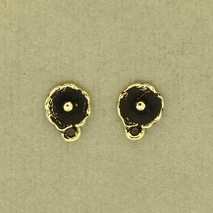 Puede incluir: Dos pequeños dijes de joyería en forma de flor con un borde festoneado en tono dorado y un centro oscuro hundido con una cuenta central dorada. Cada dije incluye un pequeño lazo circular en la parte inferior para sujetarlo a una cadena.