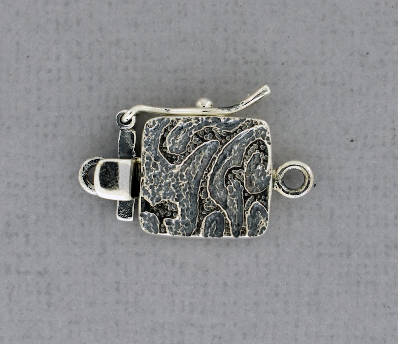 May include: Silver clasp with a textured, square design. The clasp has a loop on one side and a hook on the other.