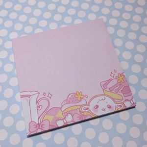 May include: A pink notepad with a decorative border featuring a cartoon design of a teapot, cupcakes, and a cute character. The illustrations are in shades of pink and yellow, with sparkling accents.