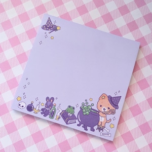 May include: Purple notepad with a witchy theme. The notepad has a purple background with a white border. The bottom border features a cartoon dog wearing a witch hat, a cauldron with green potion, a frog, a book, a bottle, a rabbit, a candle, and a skull. The top border features a witch hat with a star and a wand.