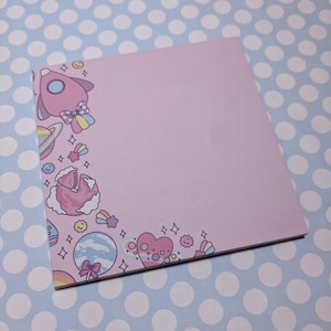May include: A square, pale pink notepad with a space-themed border. The border includes a pink rocket, planets, stars, and a heart. The notepad is set against a blue background with white polka dots.