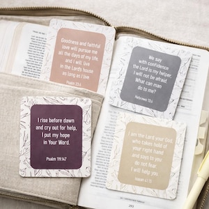 May include: Four square cards with inspirational quotes from the Bible, in a variety of colors, are displayed inside an open Bible. The cards have floral patterns and text from Psalms, Hebrews, and Isaiah. A pen rests on the open pages.
