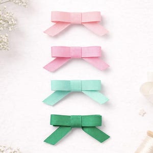 May include: Four small, colorful bows in a row. Two are pink, one is mint green, and one is dark green. The bows are made of ribbon and have a classic bow shape. The background is white with some small white flowers.