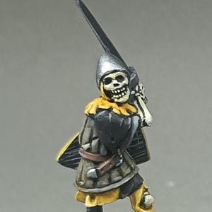 Pode incluir: Miniatura de guerreiro esqueleto, pintada com um capacete prateado, armadura preta e amarela e uma espada. A figura é montada numa base texturizada com relva verde.