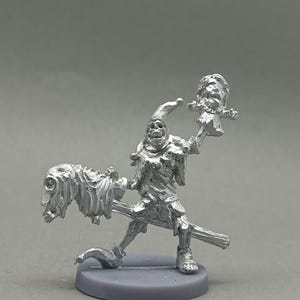 Może przedstawiać: Srebrna miniaturowa figurka błazna trzymającego dwie odcięte głowy. Błazen pozuje jakby jechał na miotle. Figurka stoi na szarej podstawie. Obrazek zawiera tekst "WolfbaneArt" w prawym dolnym rogu.