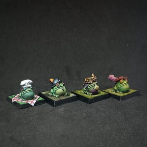 Könnte beinhalten: Vier Miniatur-Froschfiguren, jede mit einzigartiger Kleidung, stehen auf einzelnen quadratischen Sockeln. Ein Frosch trägt eine Kochmütze, ein anderer einen Hut mit Feder, ein dritter eine Krone und der letzte einen Hut mit Feder. Die Frösche sind grün mit verschiedenen bemalten Details.