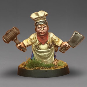 Könnte beinhalten: Miniaturfigur eines Kochs, der einen Hammer in der einen und ein Hackmesser in der anderen Hand hält. Der Koch trägt eine weiße Schürze, eine Kochmütze und ein rotes Halstuch. Die Figur steht auf einem kleinen, runden Sockel mit Gras.