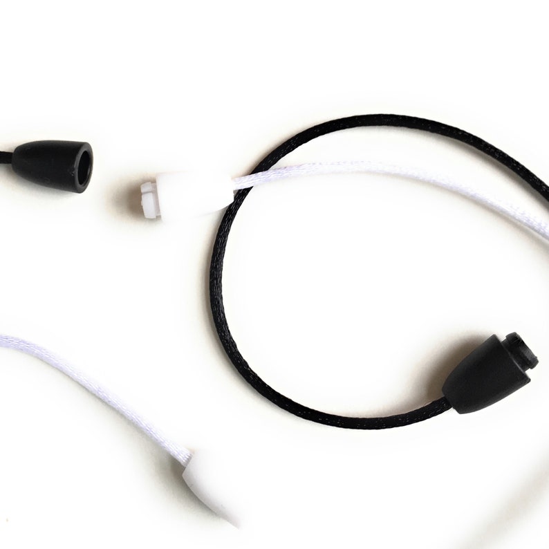 May include: Close-up of black and white drawstrings with black plastic cord locks. The drawstrings are made of woven material. One drawstring is black, and the other is white. The cord locks are cylindrical.