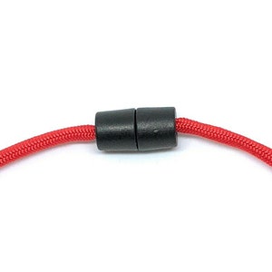 May include: Close-up of a red braided cord with a black magnetic clasp. The clasp is cylindrical and connects the two ends of the cord. The cord is a vibrant red color.