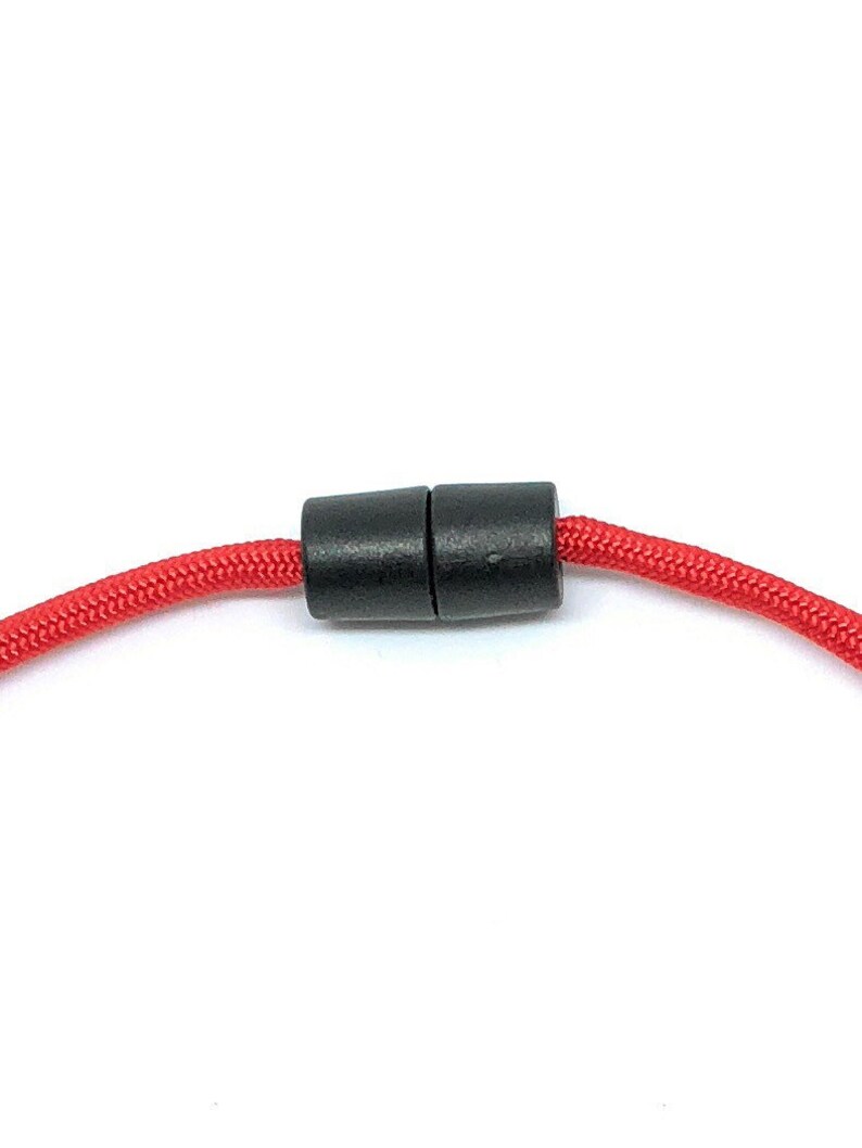 May include: Close-up of a red braided cord with a black magnetic clasp. The clasp is cylindrical and connects the two ends of the cord. The cord is a vibrant red color.
