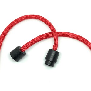 May include: Close-up of a red cord with two black plastic end pieces. The cord is thick and braided, and the end pieces are cylindrical. The background is plain white.