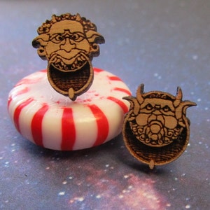 May include: A pair of wooden earrings featuring a carved design of a grotesque face with horns. The earrings are brown and have a ring-like detail around the face.