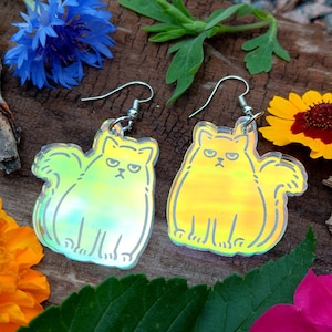 May include: A pair of iridescent cat earrings with silver hooks. The cats are facing forward with a grumpy expression. The earrings are on a wooden surface with flowers.