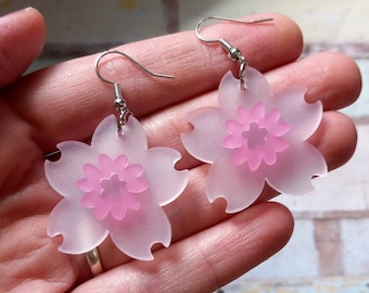 May include: A pair of pink frosted acrylic earrings shaped like cherry blossoms. The earrings have silver hooks.