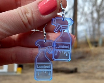 May include: A pair of translucent light blue acrylic earrings shaped like spray bottles. Each earring features a white printed label with the text Luminol and a dripping liquid graphic at the base. The earrings hang from silver-toned metal hooks.
